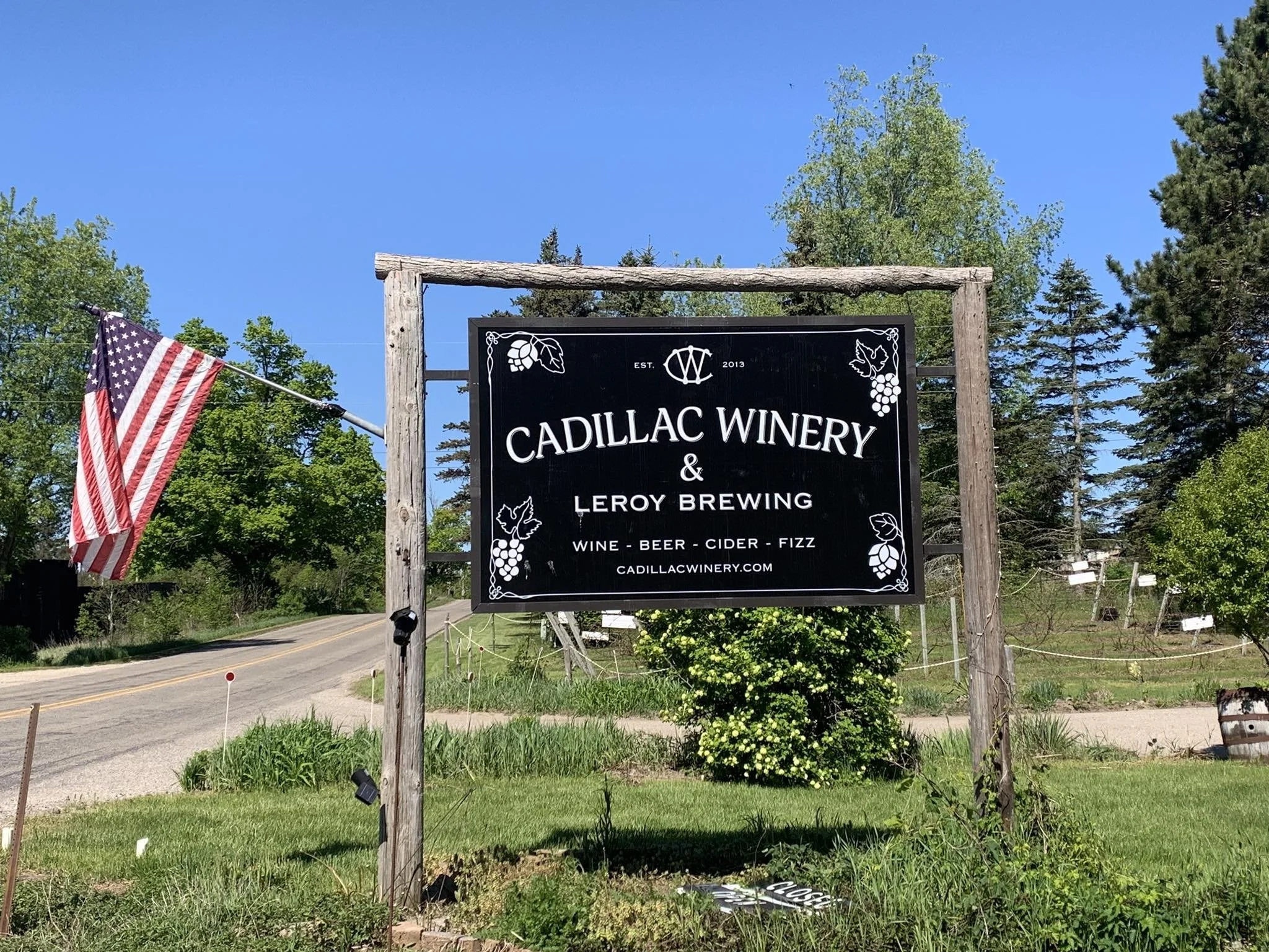 Sign for Cadillac Winery and Leroy Brewing with decorative wine and leaf graphics, an American flag on the left, and surrounded by trees and blue sky.