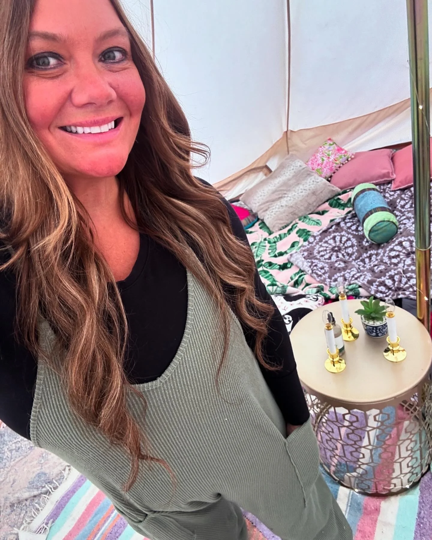 A woman with long, wavy red hair smiling for the camera in a pink and white tent, with a round table decorated with small candles and a succulent plant behind her. The table has gold candle holders and is on a colorful striped rug.