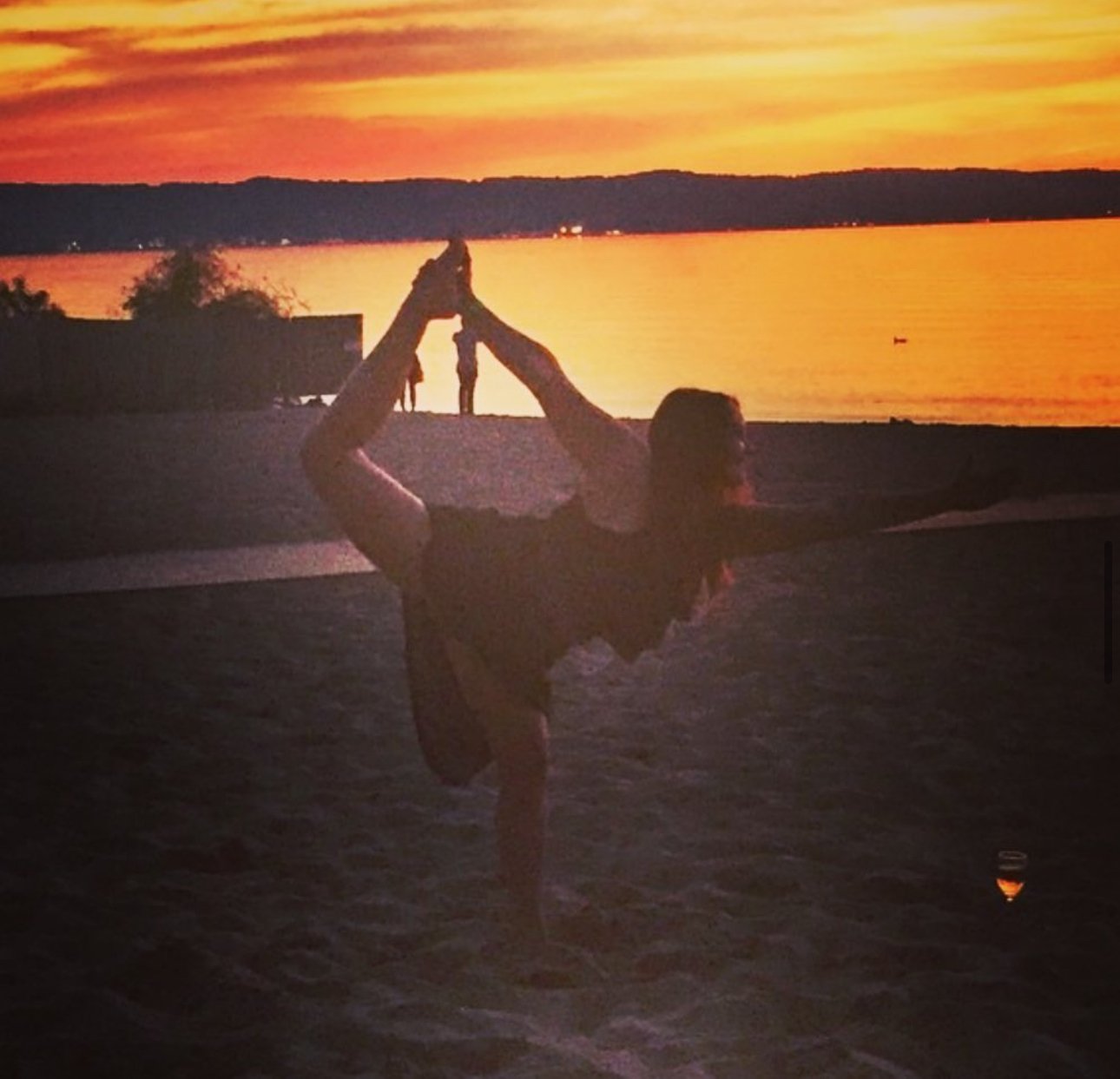 Person practicing yoga on a beach at sunset, performing a balancing pose with arms extended, holding each other's hands, with a calm water body and orange sky in the background.