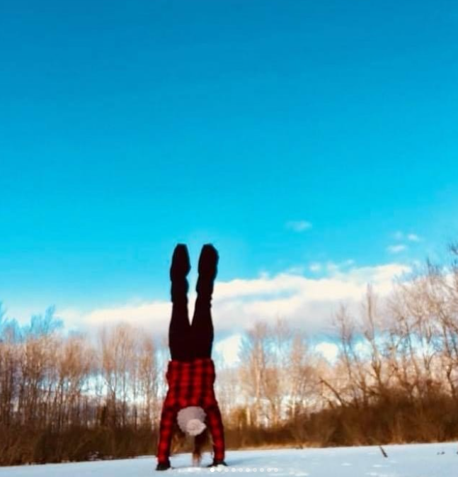 Person performing a handstand on snow-covered ground in a winter landscape with bare trees and a blue sky.