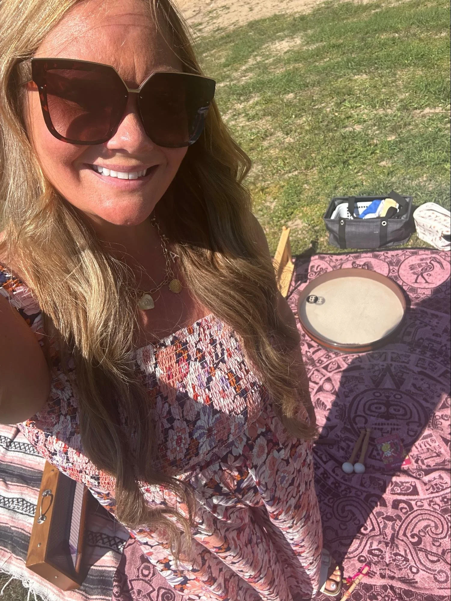 A woman with long blonde hair wearing sunglasses and a floral dress, smiling outdoors. She is sitting at a table with a patterned pink tablecloth, with various objects on the table including a circular tray, a black bag with items inside, and other s