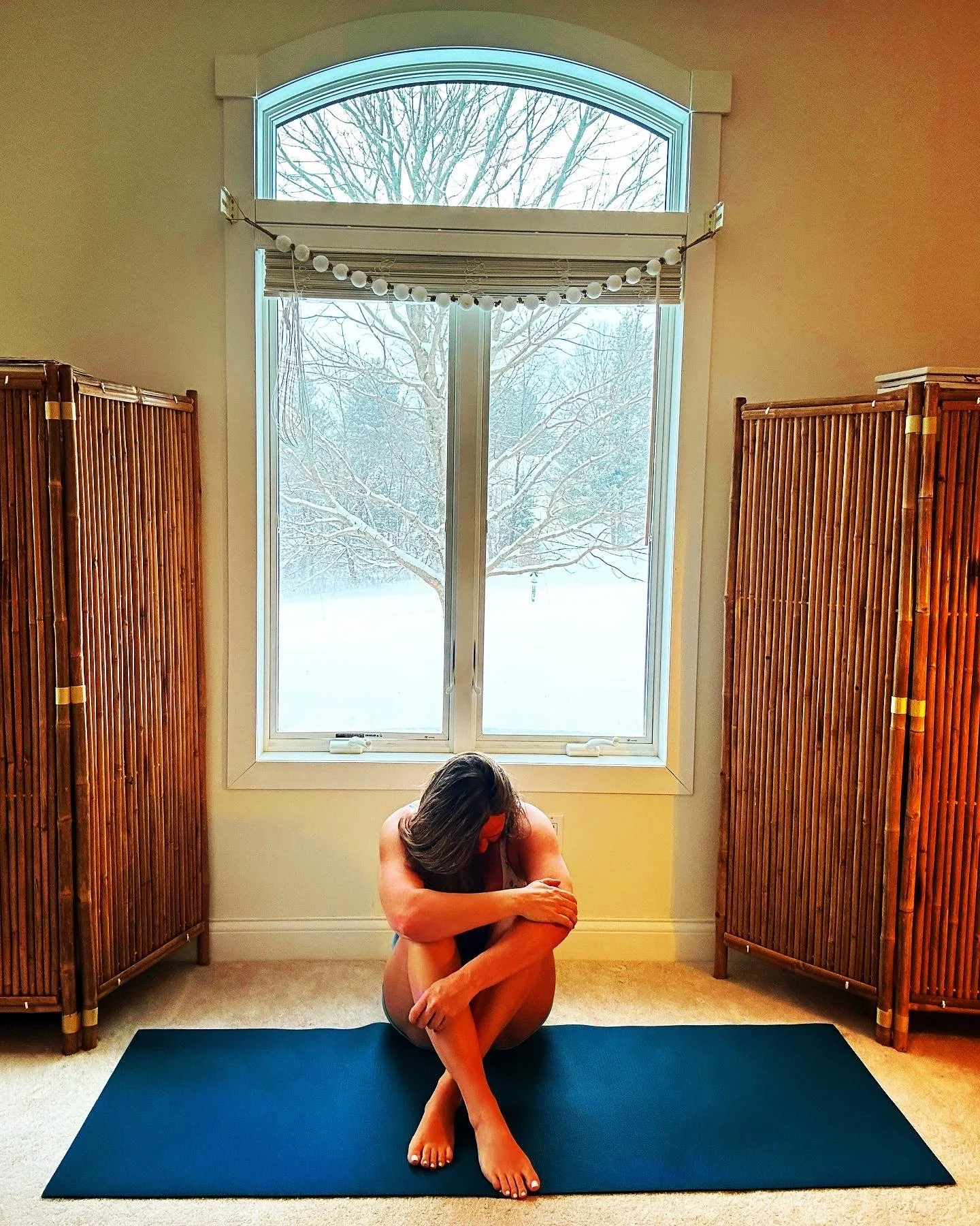 A woman practicing yoga or meditation on a black yoga mat in a room with beige carpet, flanked by two wooden room dividers, in front of a large window showing a snowy winter scene outside.