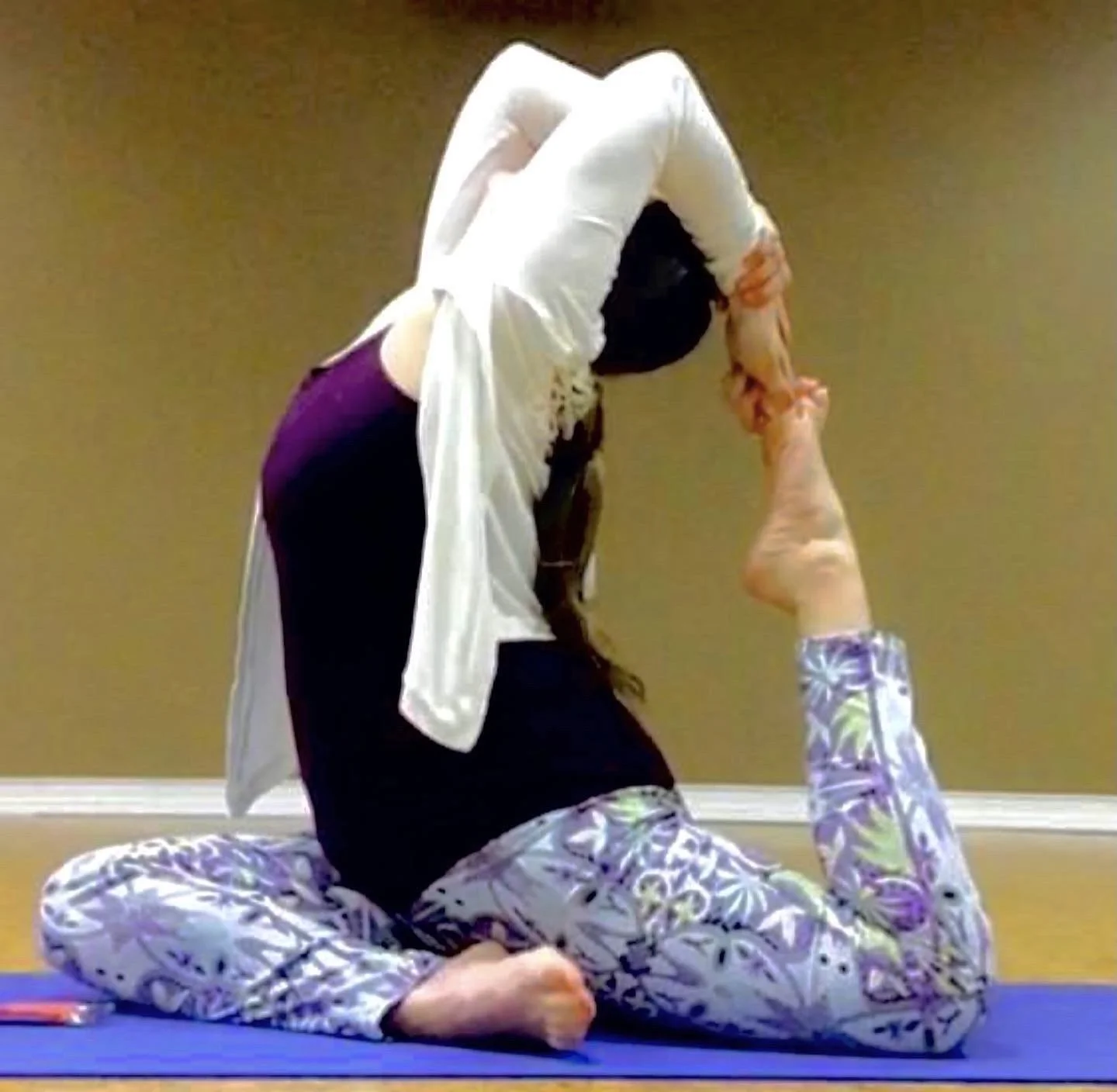 Two women practicing AcroYoga in a room with a yellow wall. One woman is lying on her back on a mat, holding the other woman who is bent over, with her head near the ground and supported by the woman on the ground.