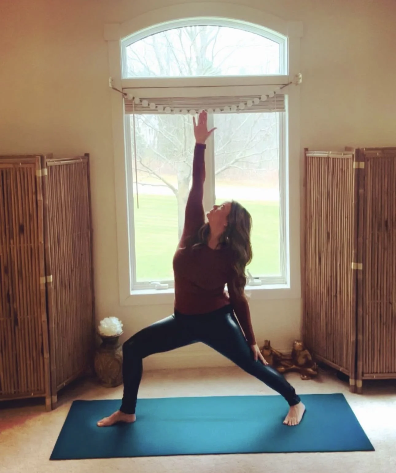 Woman practicing yoga in a spacious room with a large window, wooden room dividers, and a yoga mat on the floor.