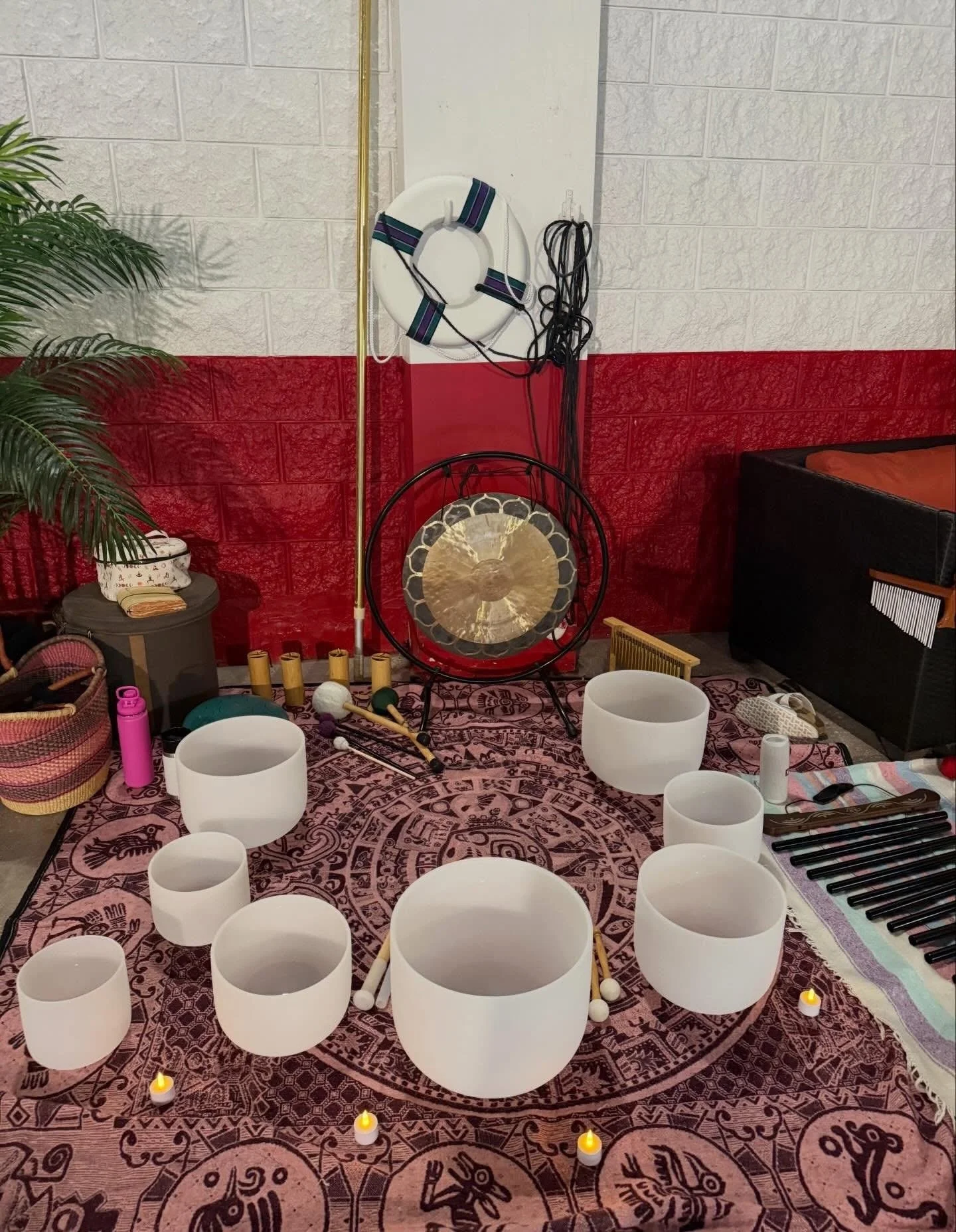A room set up for a sound healing or meditation session, with white crystal singing bowls arranged on a patterned pink and purple rug, surrounded by small candle lights. There are kundalini yoga drums, mallets, a gong, a guitar, and other musical ins