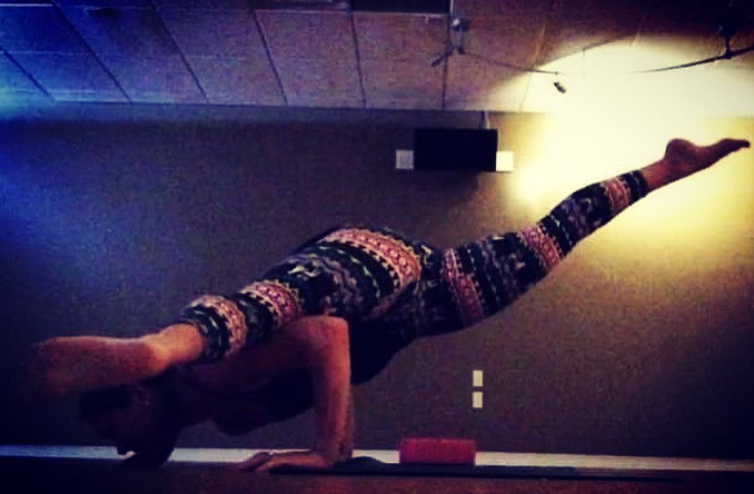 Person performing a yoga pose in a dimly lit room with a wooden ceiling, wearing colorful patterned leggings and a black top.