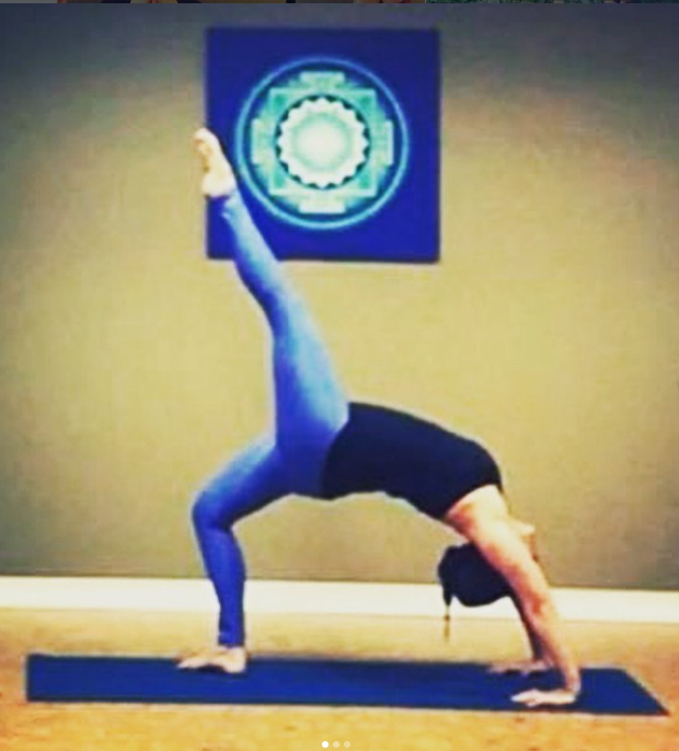 Person in a black tank top and blue leggings performing a yoga pose called Dolphin Pose in a room with a gray wall and a blue artwork with a geometric pattern hanging on the wall.