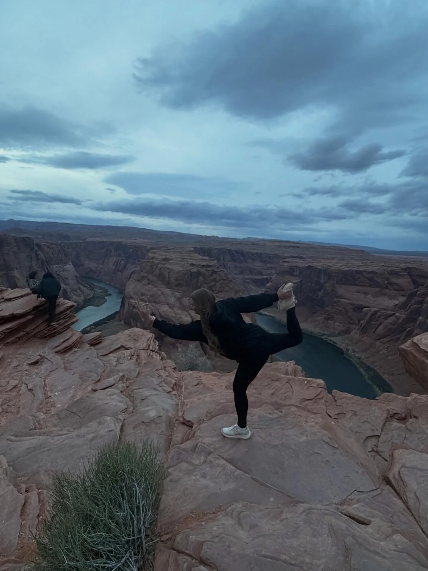 Person in black clothing doing a yoga pose on rocky ledge overlooking a canyon and river under a cloudy sky.