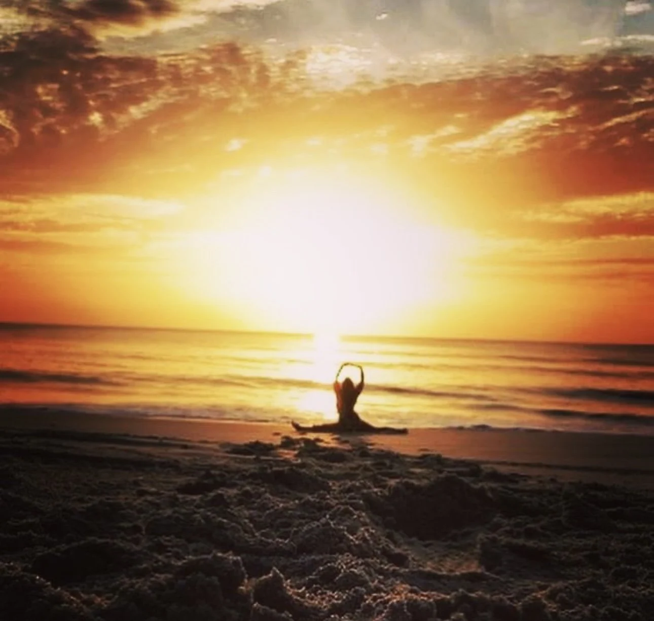 Person practicing yoga or meditation on the beach at sunset with the sun low on the horizon and the sky filled with colorful clouds.