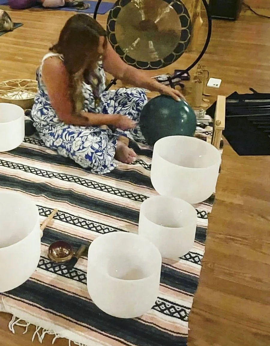 A woman seated on a striped rug, playing a gong with a mallet surrounded by crystal singing bowls in a yoga or meditation space.