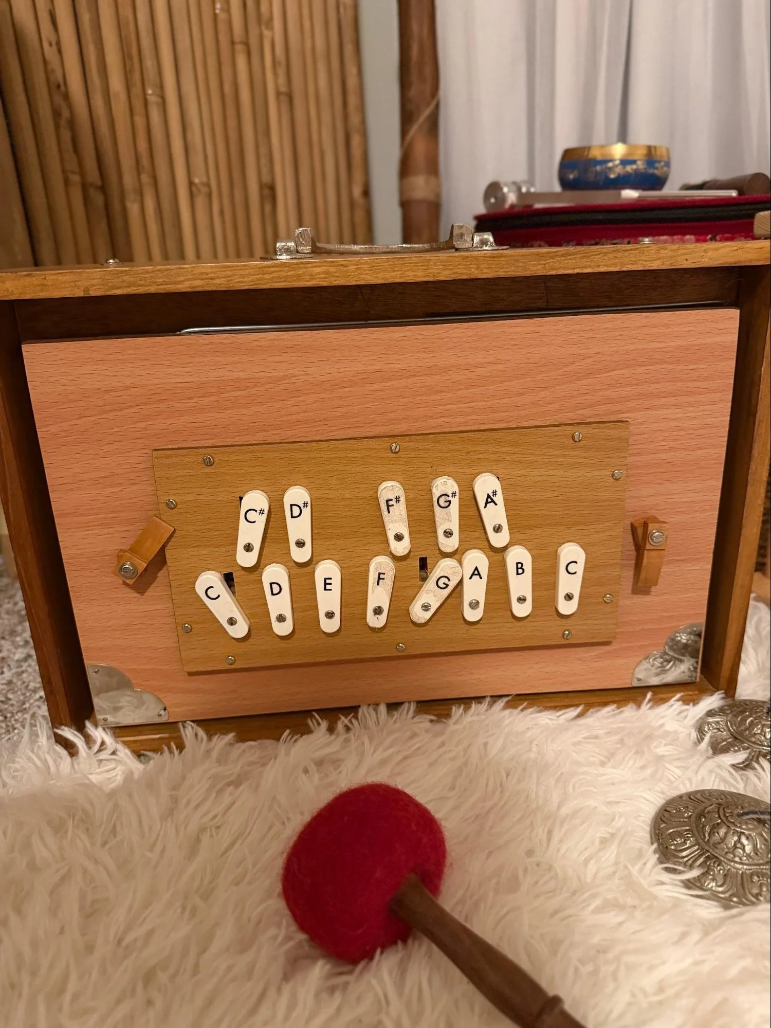 A wooden toy organ with white and black keys and labeled white buttons, placed on a fluffy white rug. There is a red mallet with a wooden handle and a red felt-covered head in front of the organ. To the right, there are decorative silver objects. The