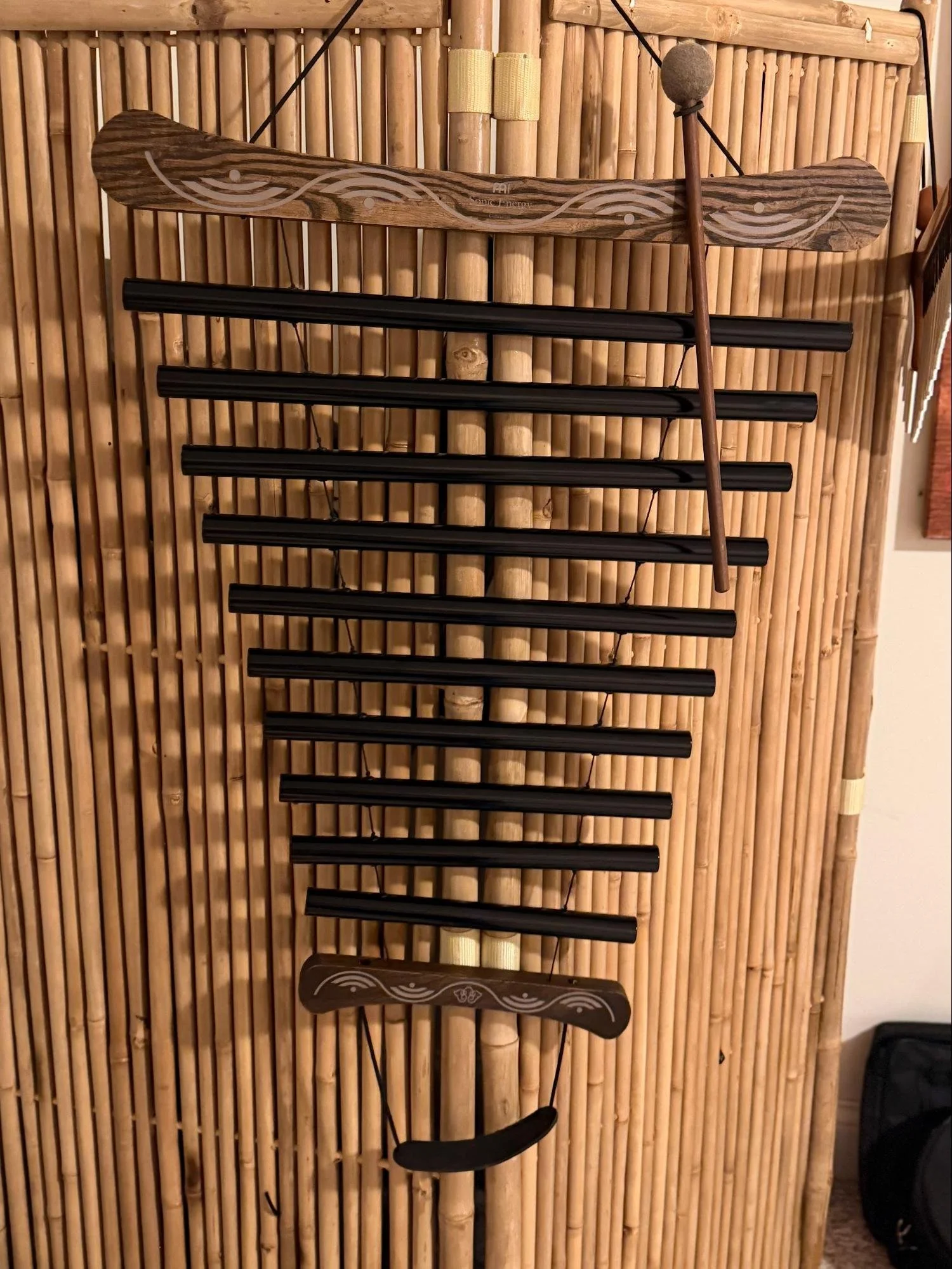 A wooden wall display with a yak horn shaped sculpture at the top, three black horizontal bars, and a wooden percussion instrument resembling a marimba at the bottom. The background is made of bamboo sticks.