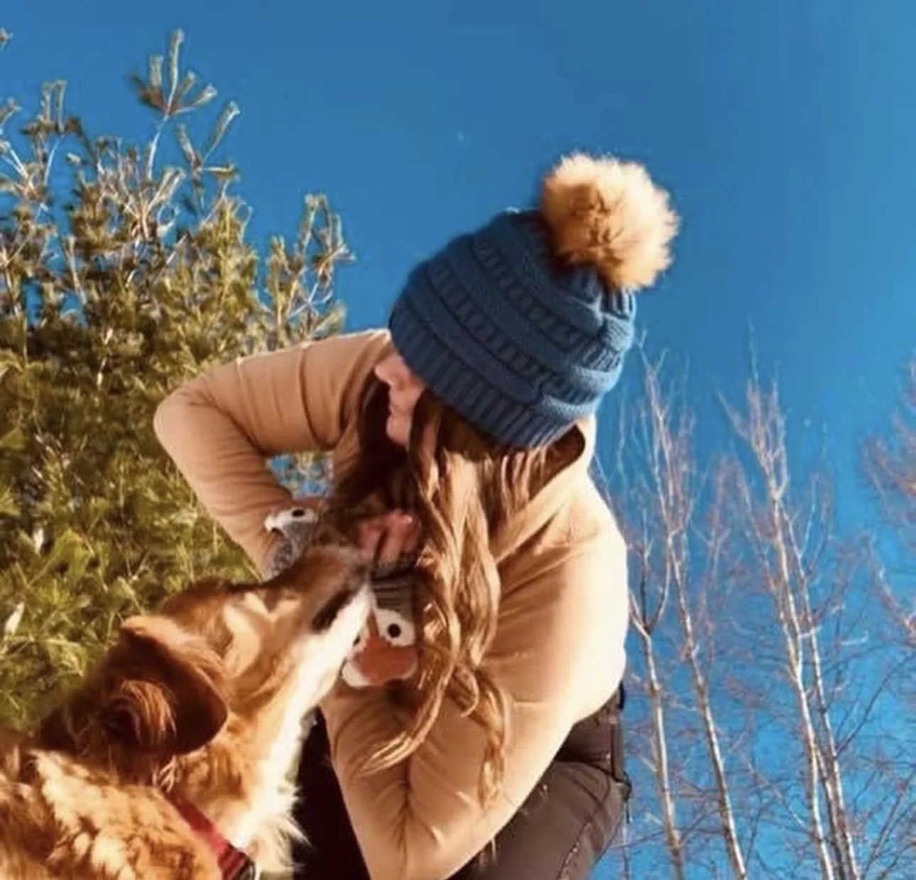 Person with long hair in a tan coat and a blue knit beanie with a pompom, outdoors among trees, holding the snout of a brown and white dog.