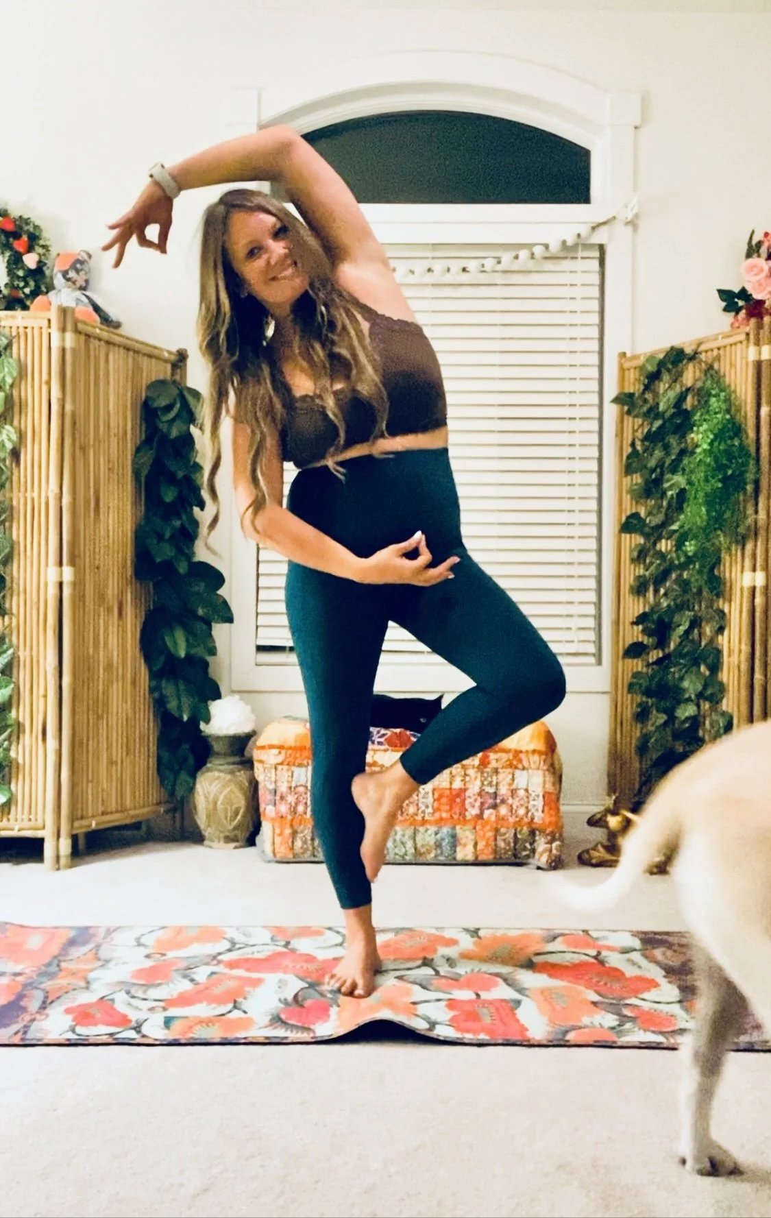 A woman practicing yoga indoors, standing on one leg on a colorful floral yoga mat, with her other leg bent and foot resting on her inner thigh. She is performing a dance pose with one arm raised above her head and the other hand holding her ankle. T