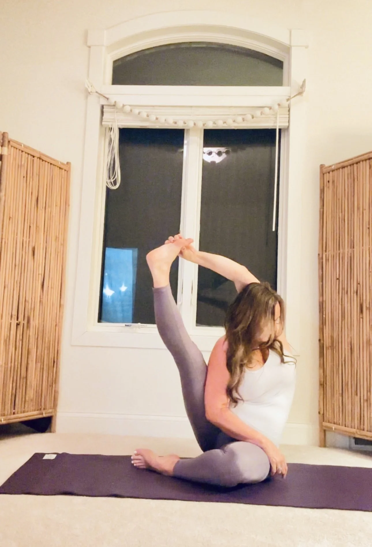 A woman practicing yoga indoors, performing a backbend with a leg extended upward, holding her foot with one hand, on a yoga mat in front of a window with bamboo blinds.