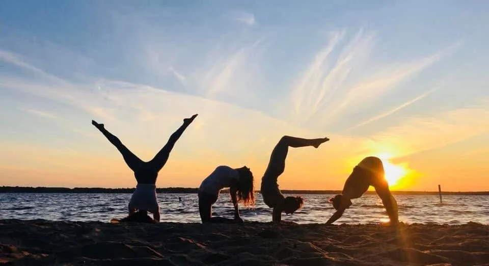 Four people practicing yoga on a sandy beach during sunset, performing various poses with the ocean in the background.