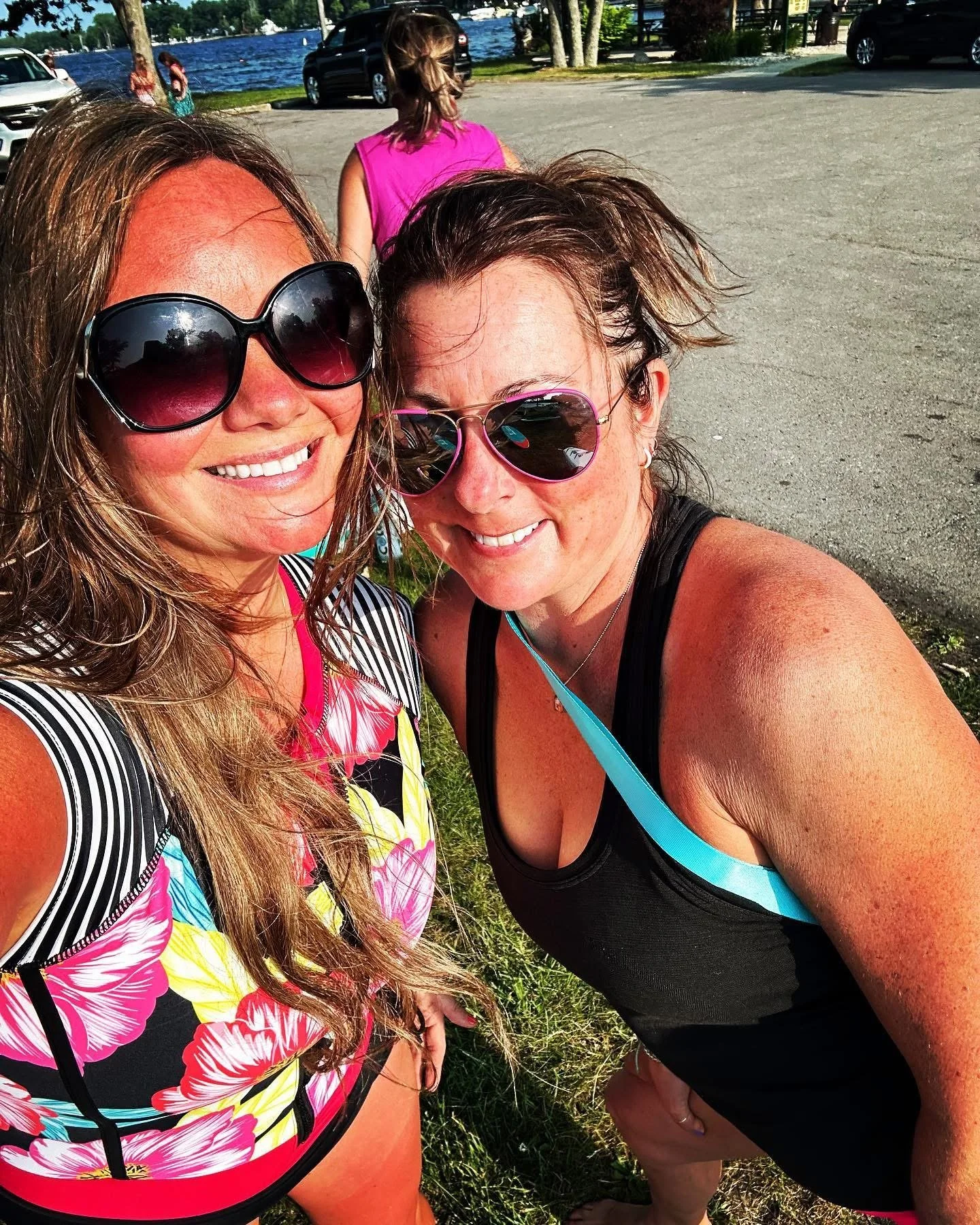Two women taking a selfie outdoors near a body of water, wearing sunglasses and casual summer clothing, with a car and trees in the background.