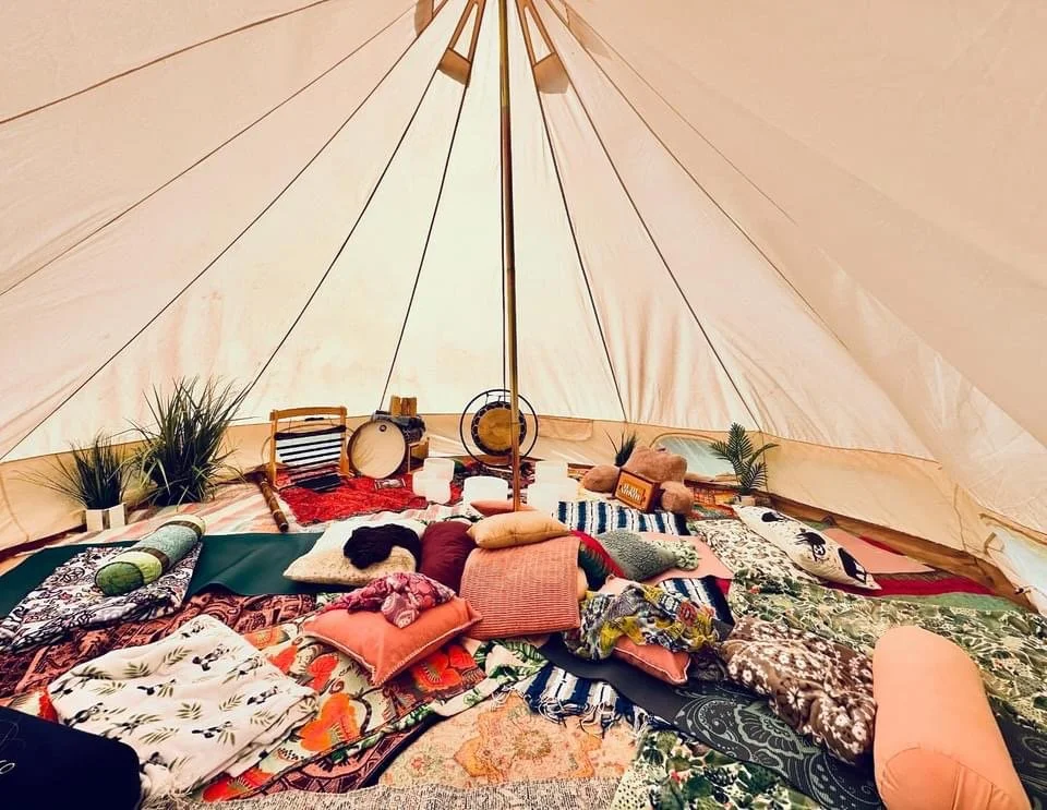 Inside a large, cozy tent decorated with pillows, rugs, and plants, with musical instruments and candles on a low table, suggesting a relaxing outdoor glamping or camping setup.