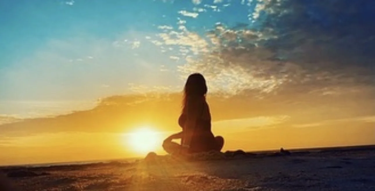 Silhouette of a person sitting cross-legged on the beach during sunset, with a colorful sky and clouds.