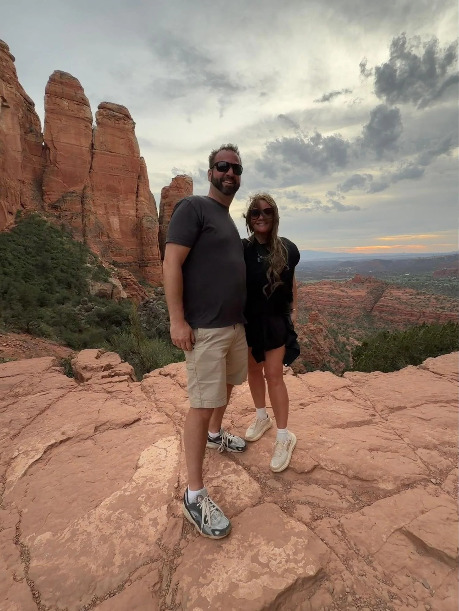 A man and woman standing on red rocks in a canyon during sunset. The man is wearing black sunglasses, a black T-shirt, beige shorts, and sneakers. The woman is wearing sunglasses, a black dress, and sneakers. There are tall red rock formations and a 