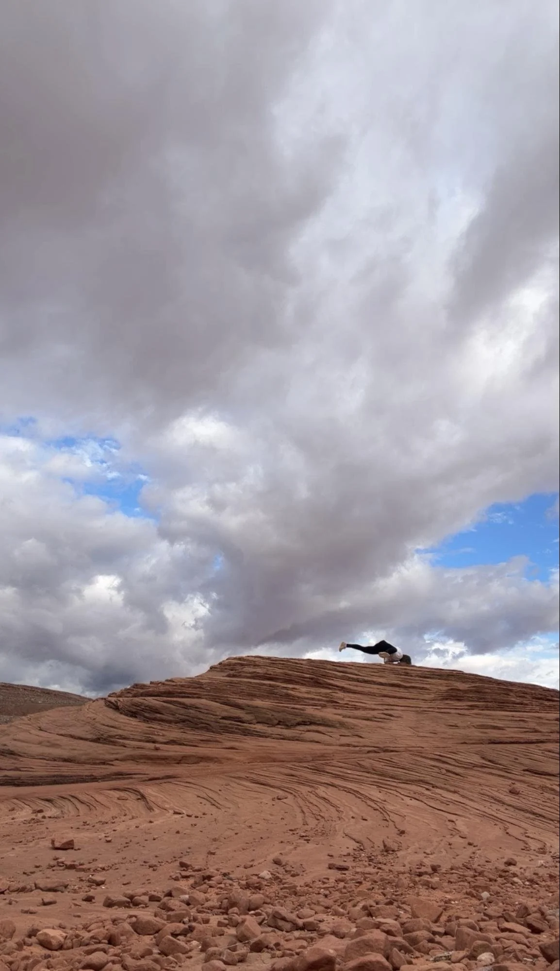 Person performing a headstand yoga pose on a rocky desert landscape with a cloudy sky in the background.
