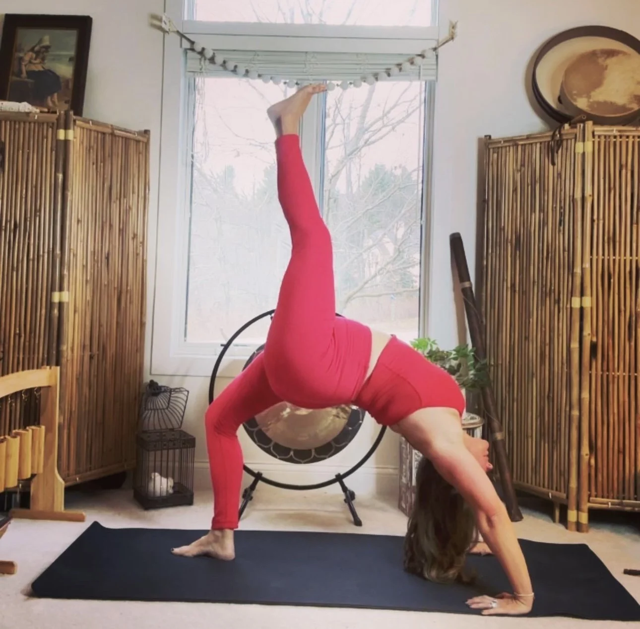 A woman wearing red leggings and a matching top practicing yoga indoors, performing a reverse wheel pose on a black yoga mat.