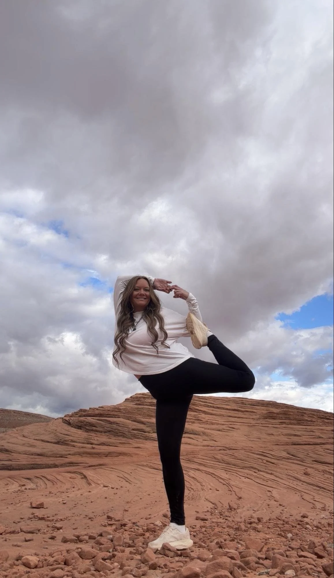 A woman performing a yoga pose outdoors on a rocky desert landscape with cloudy skies in the background.