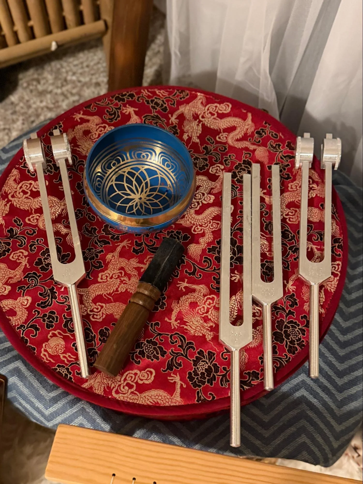 A collection of musical instruments on a red embroidered cloth, including gongs, a mallet, and a metal bowl with intricate designs, with a wooden chair and a curtain in the background.