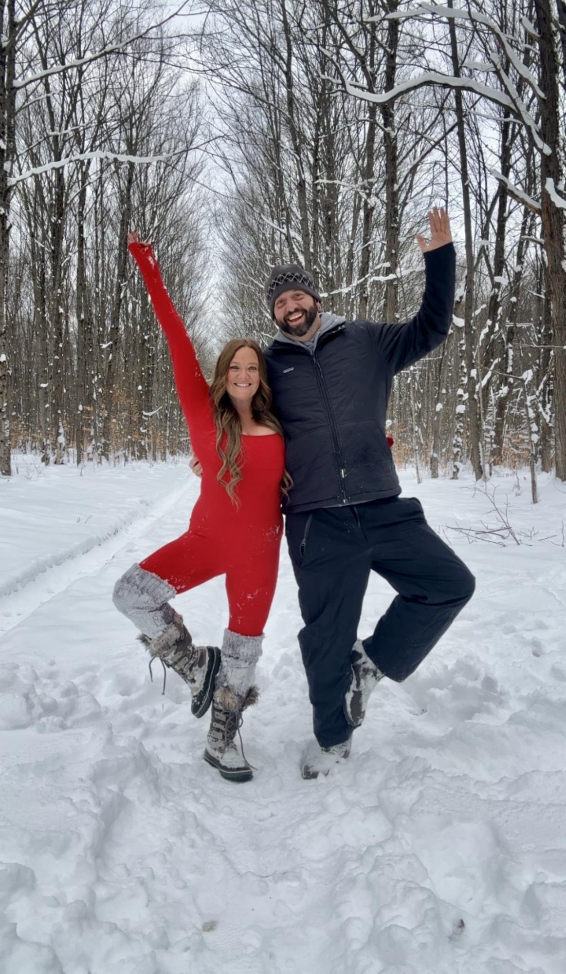 A woman in a red jumpsuit and a man in a black jacket and pants standing together in a snowy forest, both smiling and posing playfully with one leg in the air.