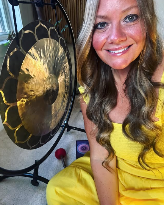 A woman with long, wavy hair wearing a yellow outfit, smiling and sitting next to a large gong with gold accents in a room with wooden walls and a window.