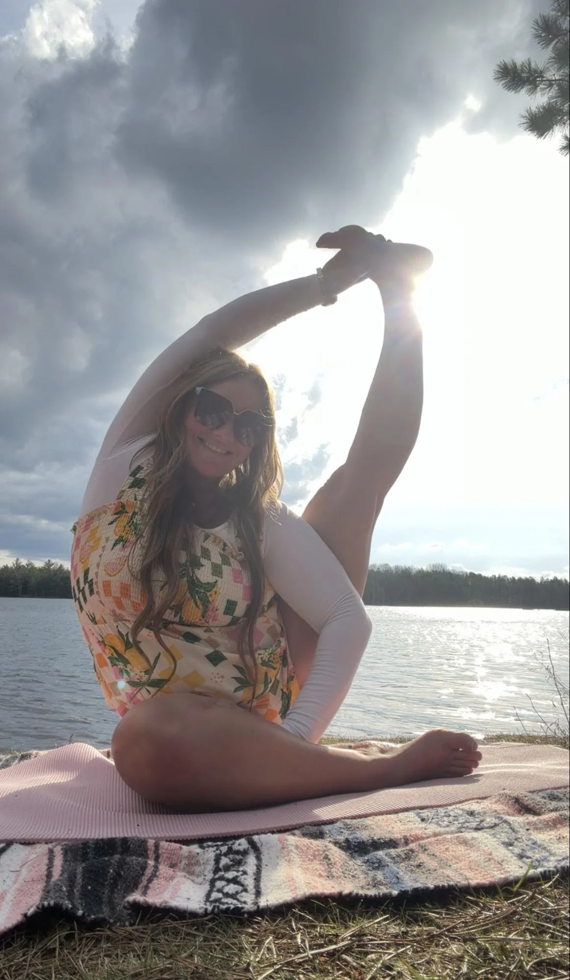 Woman in floral dress doing yoga outdoors by water, stretching her arms upward, backlit by the sun with cloudy sky and trees in background.