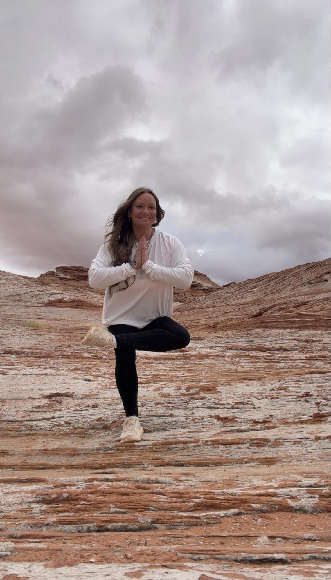 A woman practicing yoga outdoors on a rocky desert landscape under a cloudy sky, balancing on one leg with palms together in prayer position.