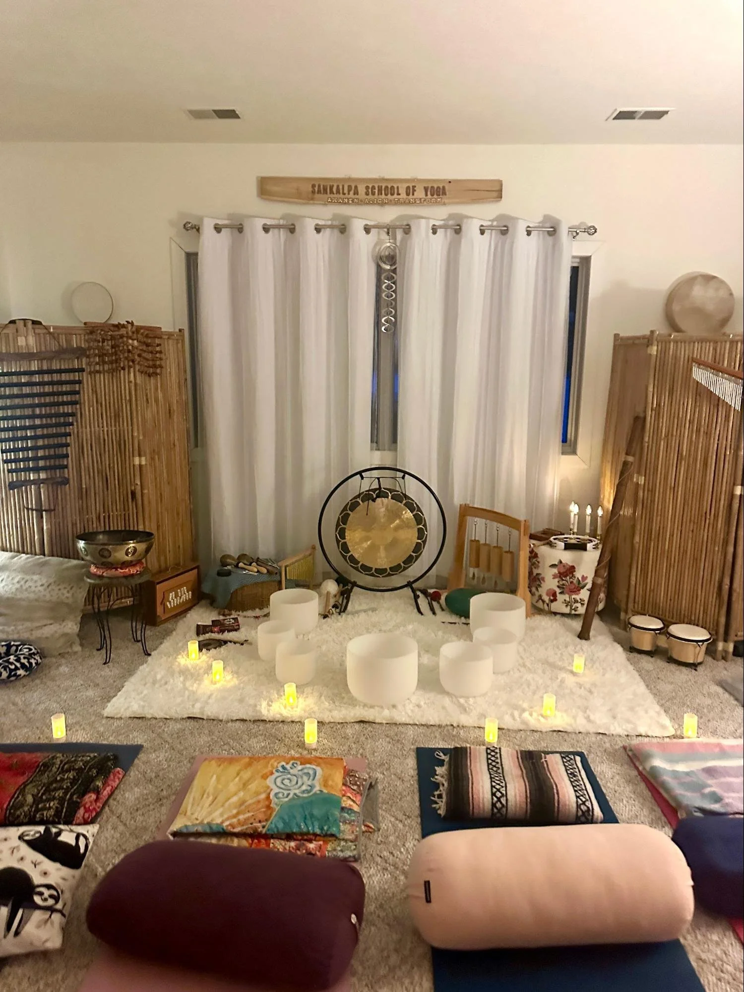 A cozy room set up for meditation or yoga, with a white shaggy rug, several candles, crystal singing bowls, and various musical and meditation instruments. There are bamboo screens on the sides, a gong in the center, and white curtains covering the w