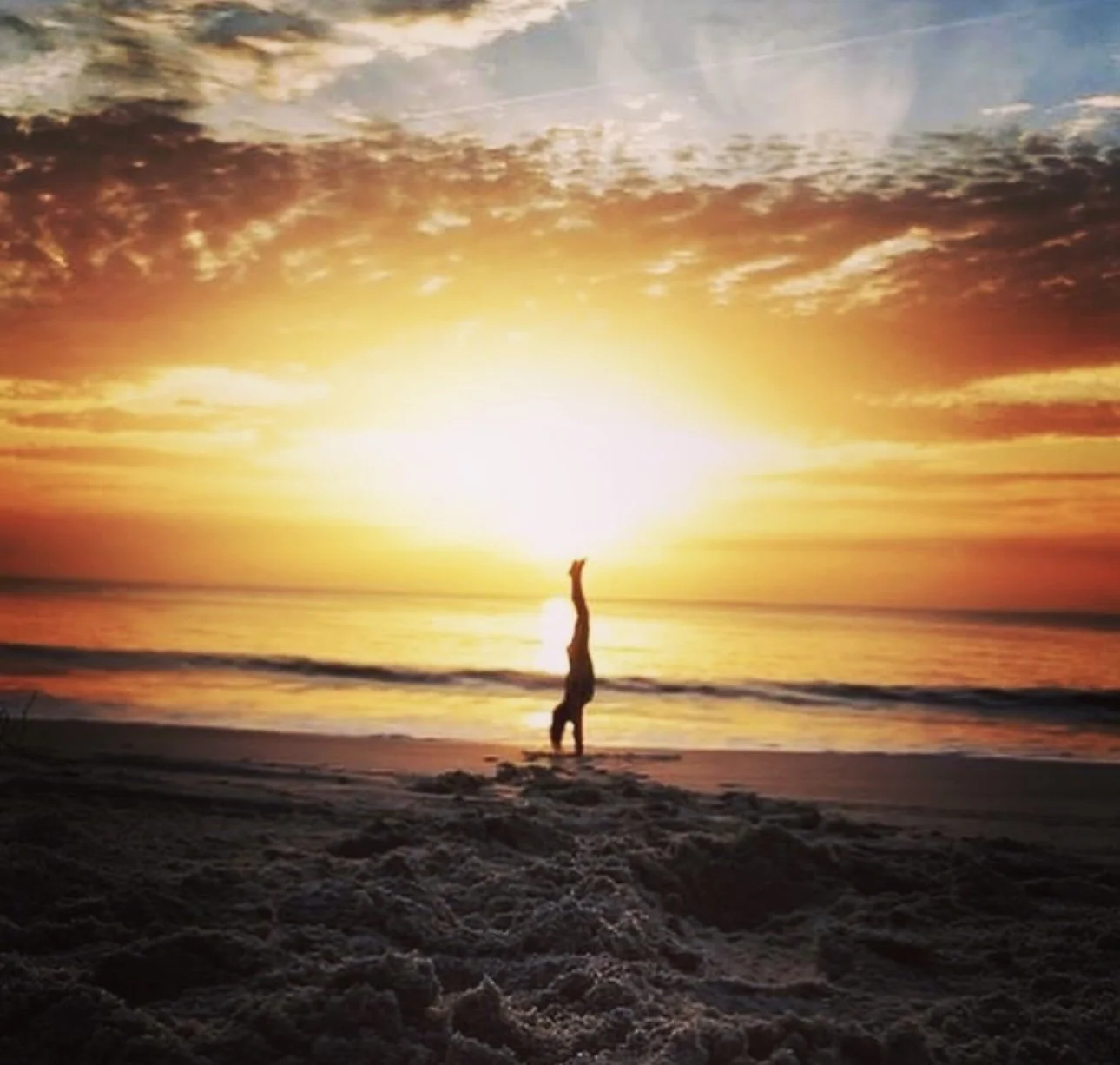 Person standing on the beach at sunset with one hand raised, silhouetted against a vibrant orange and yellow sky and reflecting sunlight on the water.