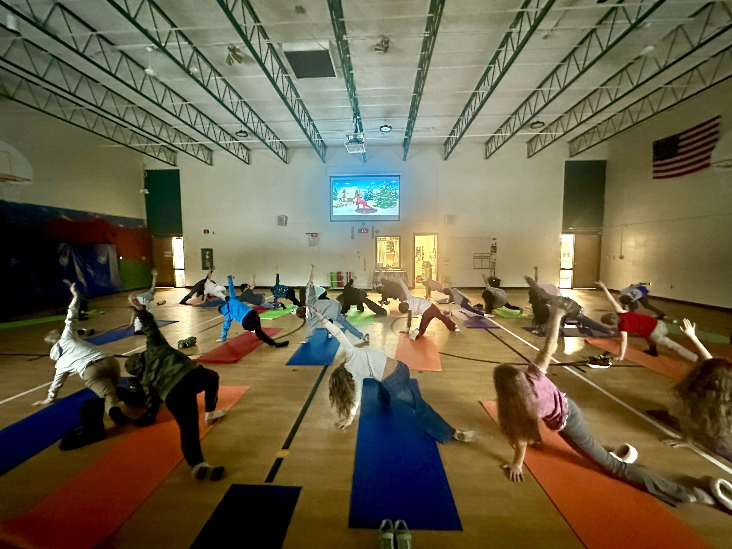 A group of children and adults participating in a yoga or stretching class in a gymnasium. They are on colorful mats, in various stretching poses, with a large screen displaying a winter scene with trees and a girl in the background.