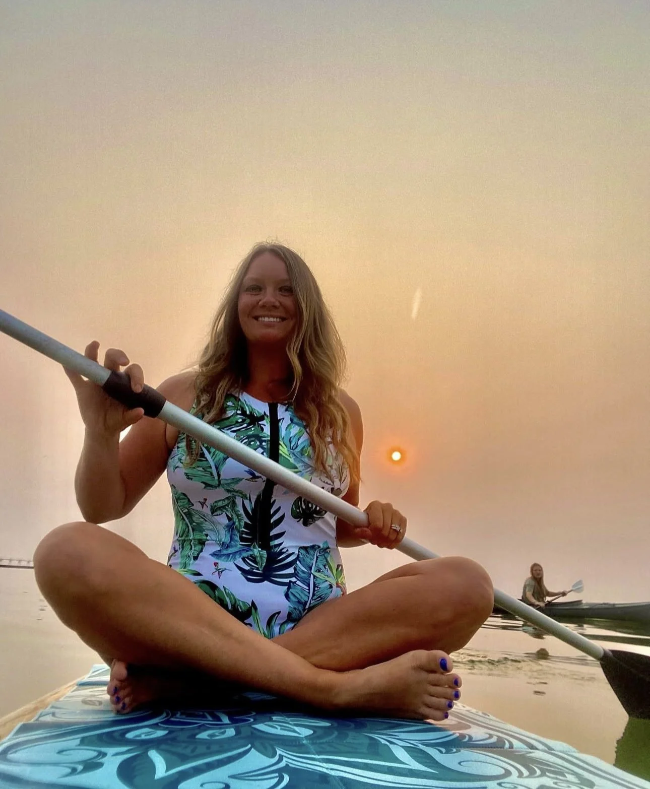 Woman sitting cross-legged on a paddleboard during sunset, smiling and holding a paddle. She wears a colorful, tropical-patterned swimsuit. In the background, a person is kayaking, and the sky has a warm hue with the sun low on the horizon.