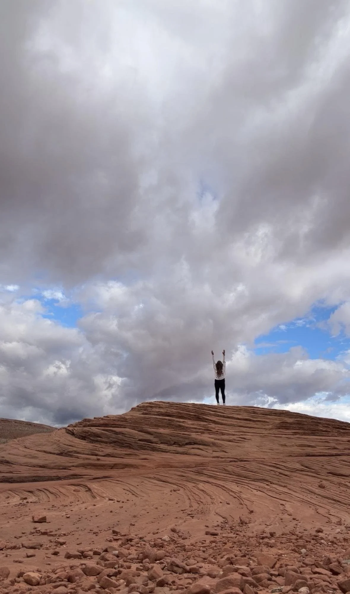 Person standing on red rock formation with arms raised under cloudy sky.