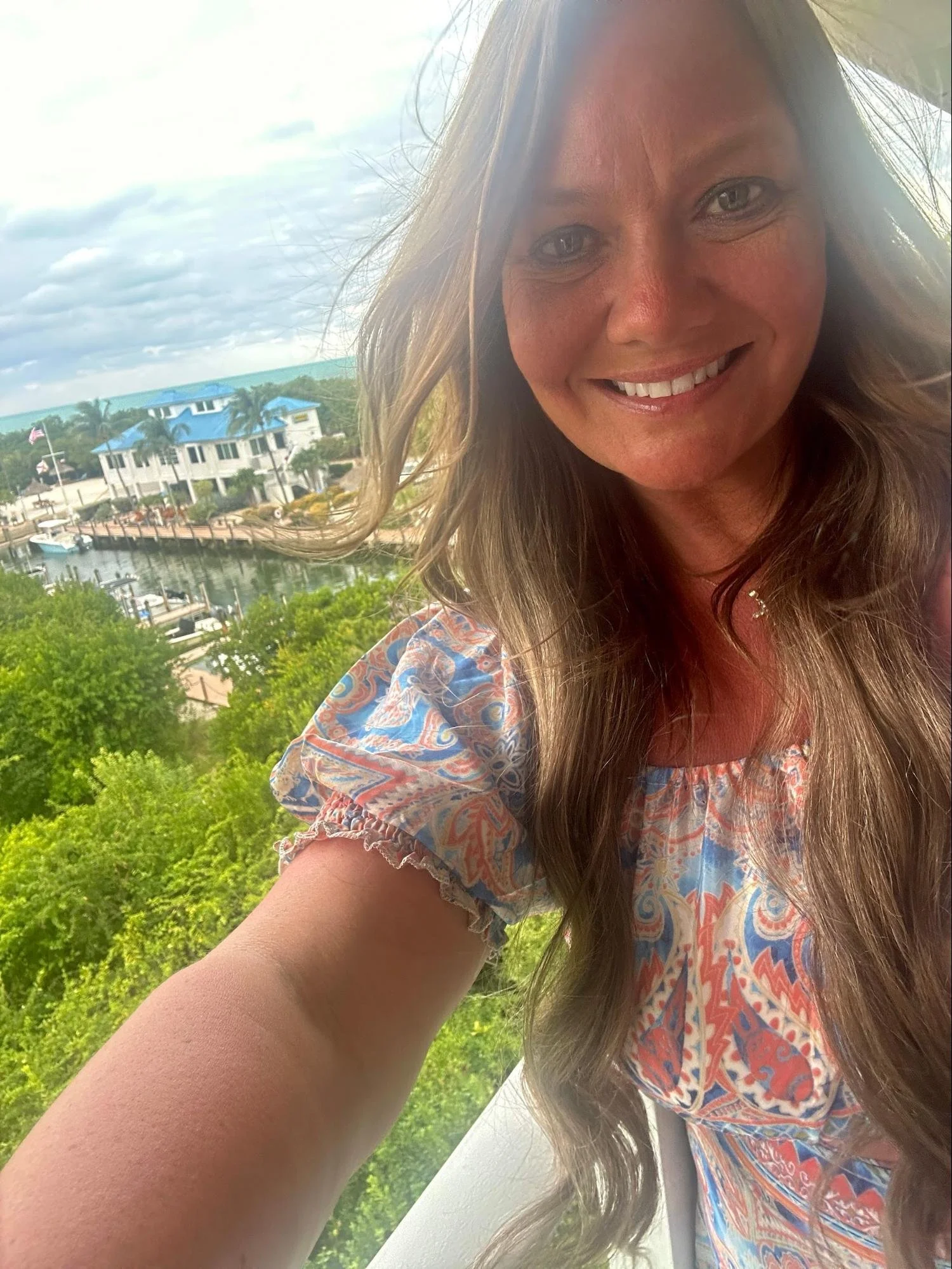 Smiling woman with long light brown hair, wearing a colorful patterned top, outdoors near water with boats and houses, cloudy sky.