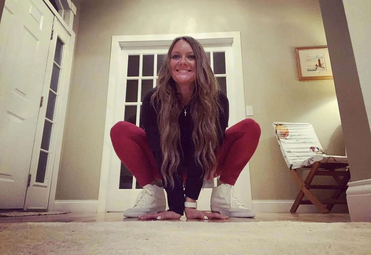 A woman with long wavy hair, wearing a black top, red pants, and white sneakers, squatting on the floor in a room with beige walls, a door with glass panes behind her, and a folding chair with colorful fabric to the right.