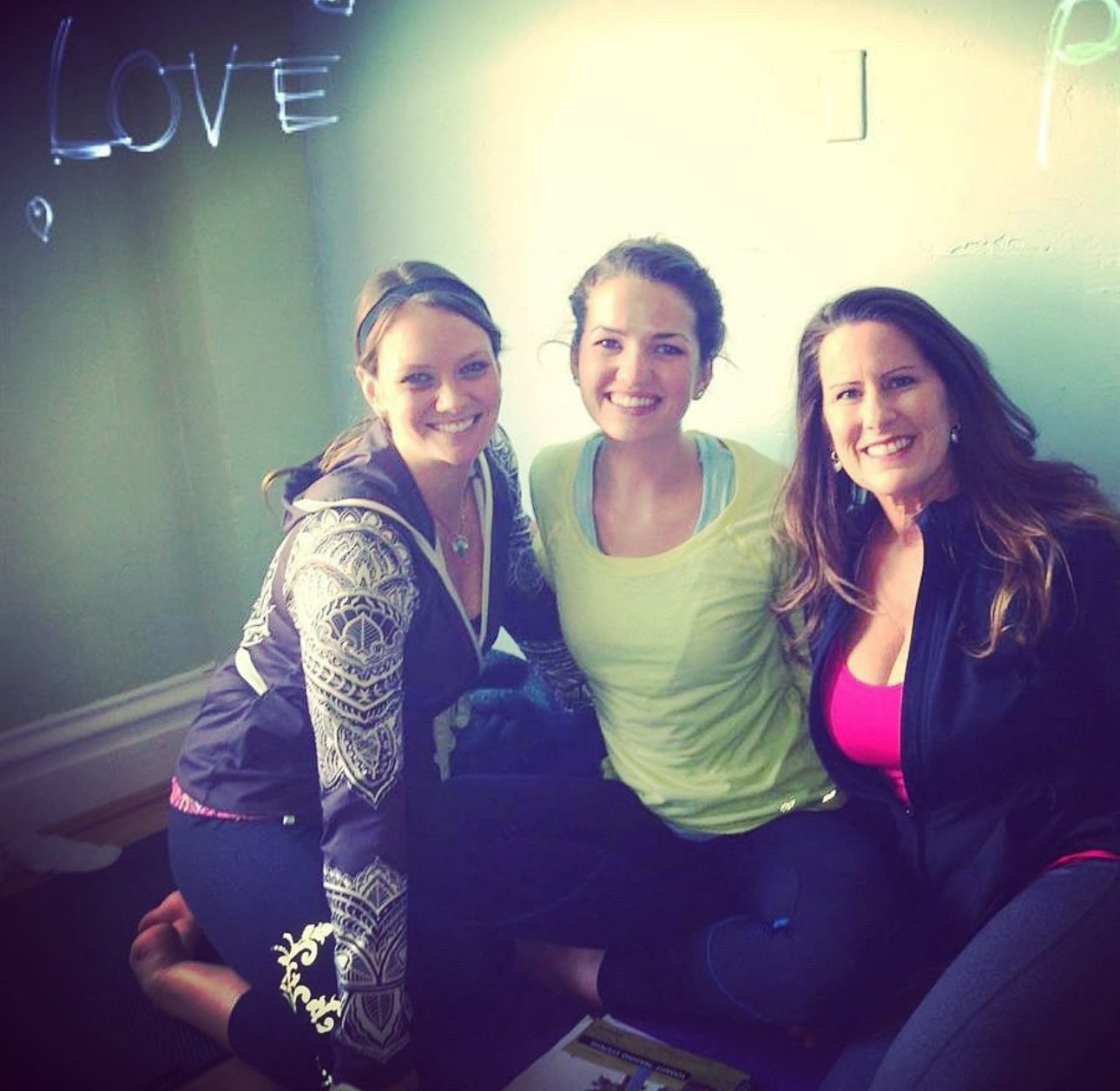Three women sitting on the floor smiling at the camera, with a green wall behind them that has the word 'LOVE' written on it.
