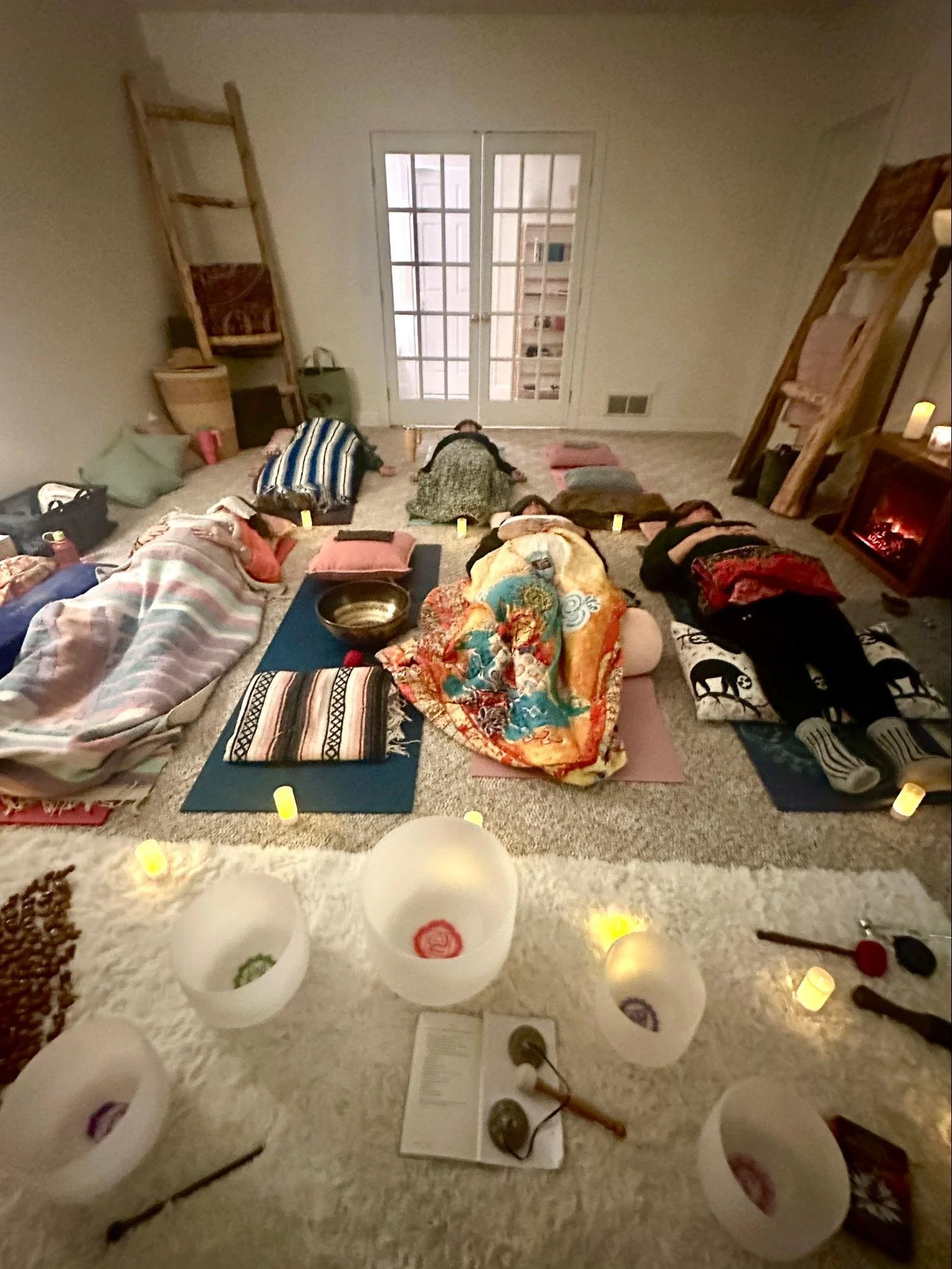 People lying prostrate on the floor with cushions, arranged in a circle, in a cozy room with a door, small bookshelf, and a fireplace.