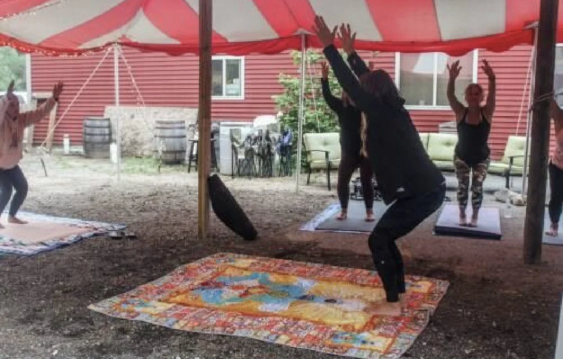 People participating in an outdoor yoga session under a red and white striped tent, with mats on the ground and a red building in the background.