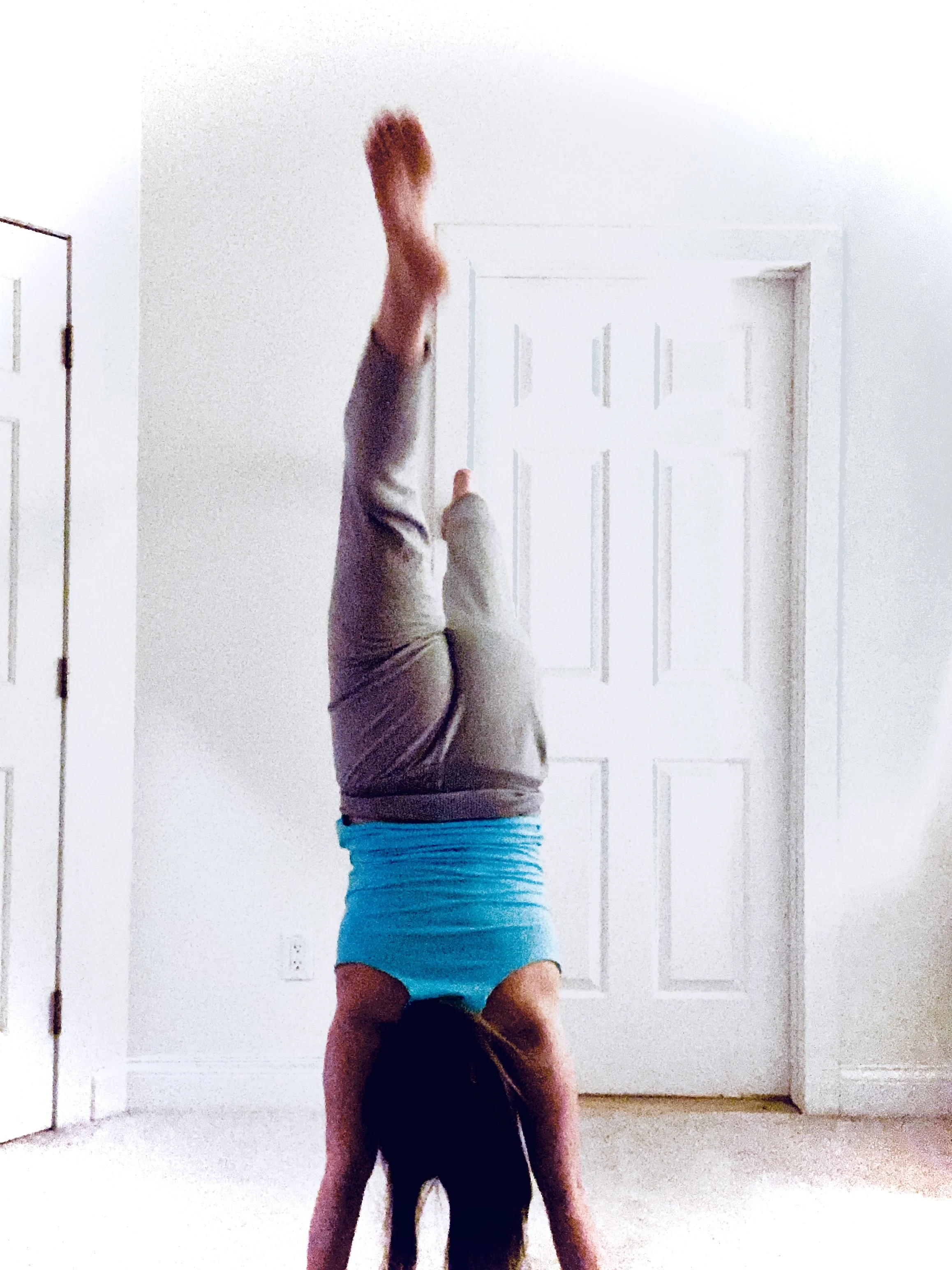 Two people perform a handstand against a plain white wall in a room with beige carpet, with one supporting the other.