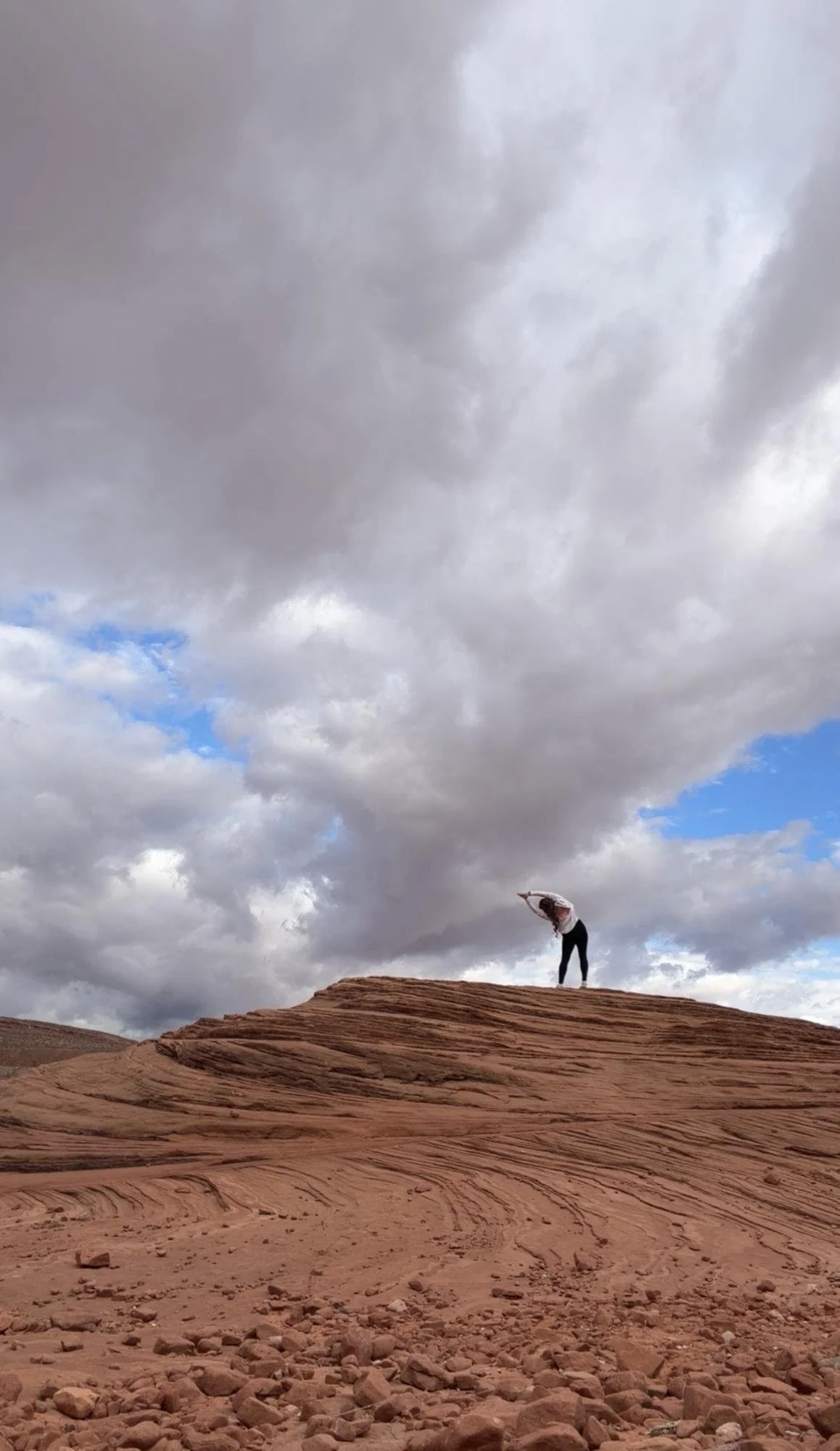 Person standing on a rocky desert landscape under a cloudy sky, stretching or reaching upward with arms extended.