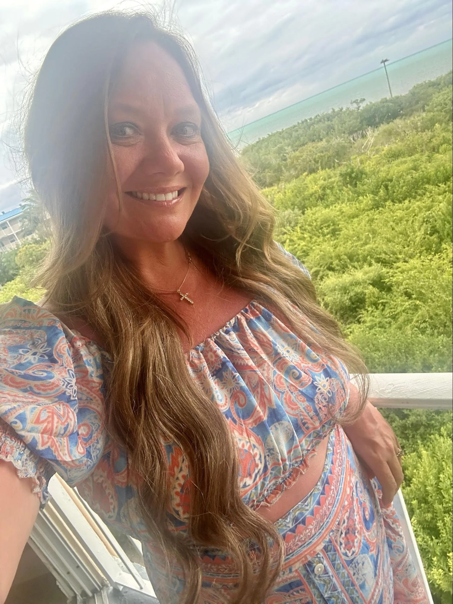 A woman with long, wavy hair smiling for a photo outdoors with a green landscape, blue water, and cloudy sky in the background.