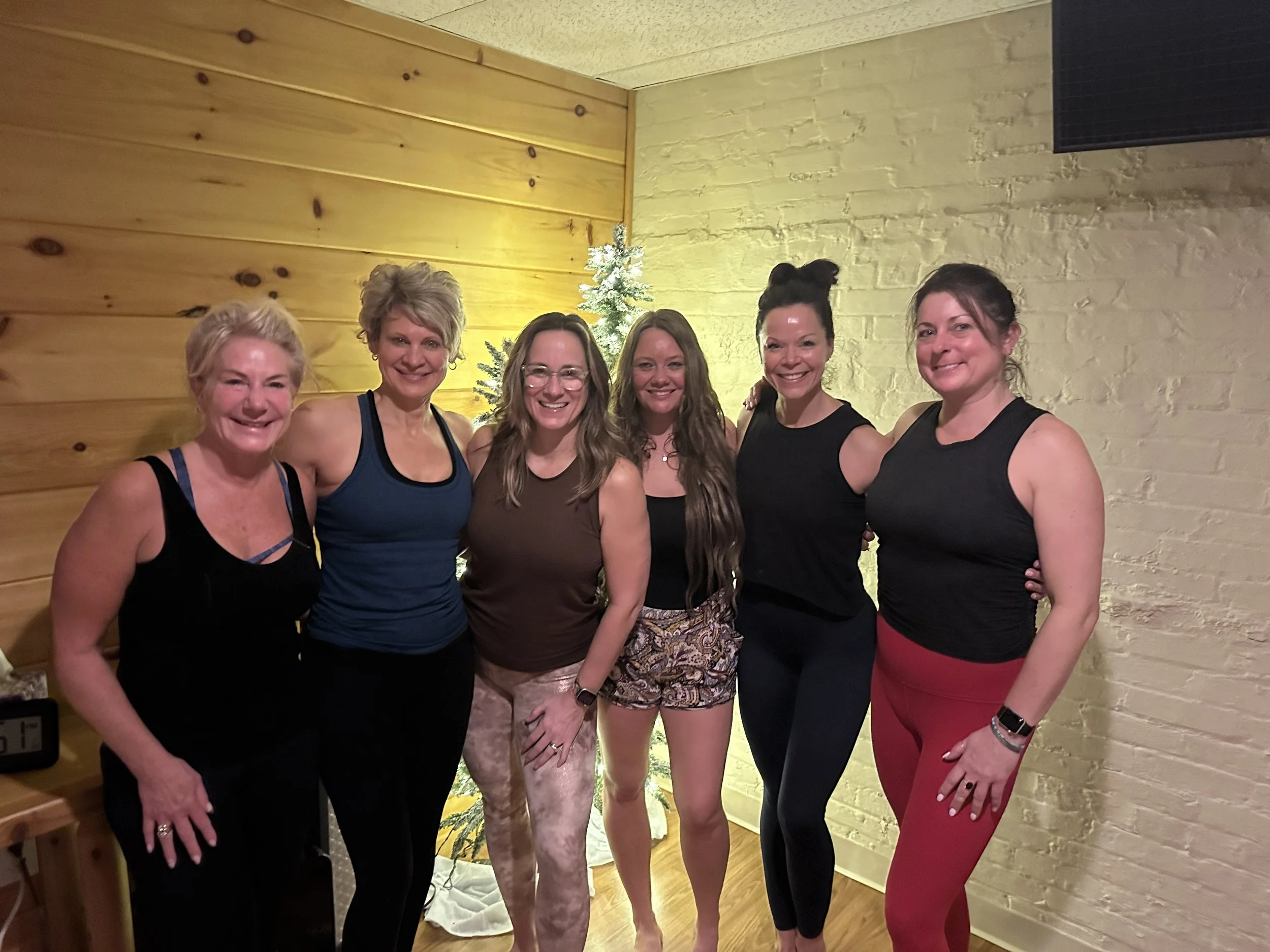 Six women standing together indoors by a small Christmas tree, smiling at the camera.