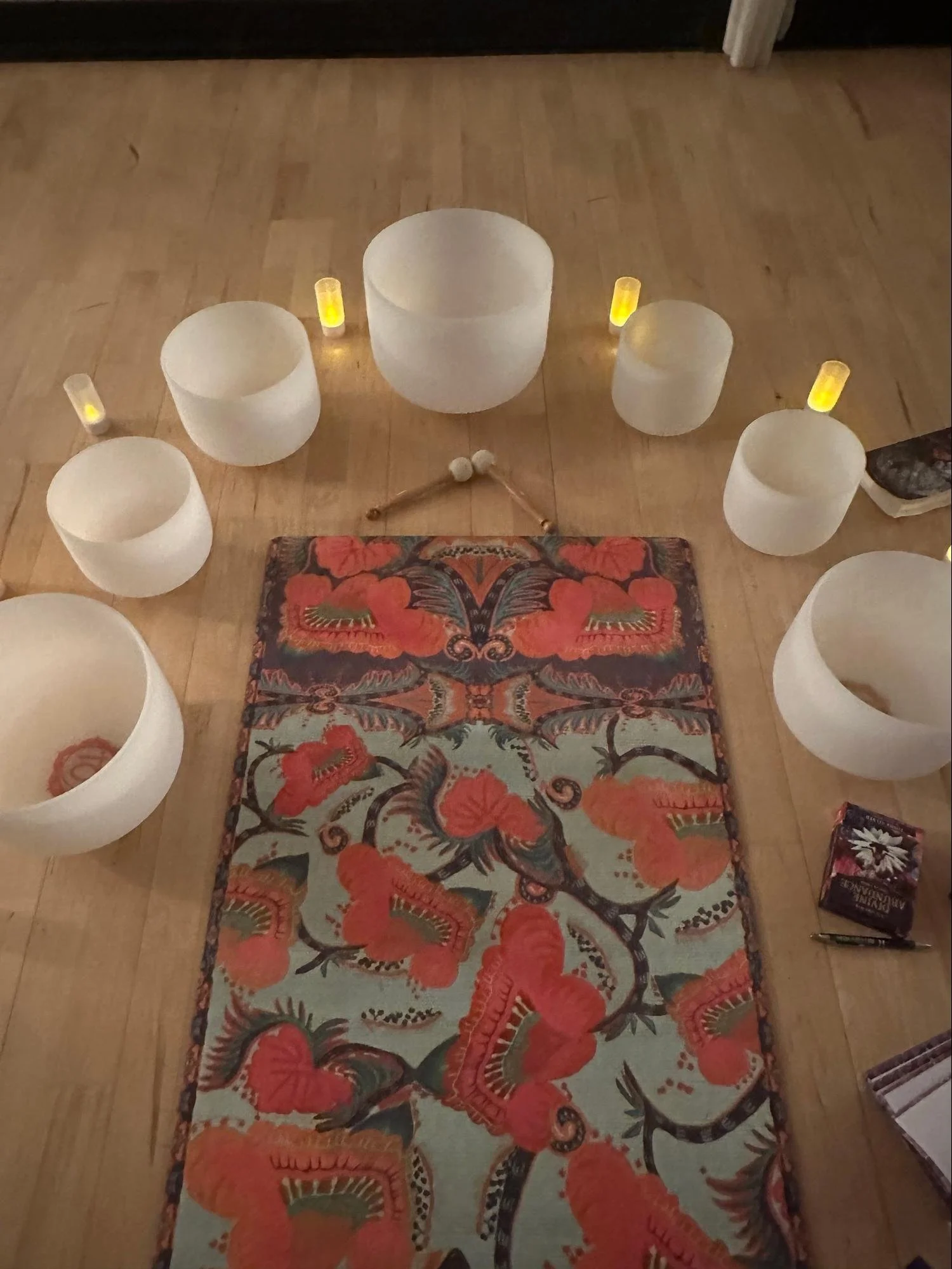A wooden floor with a colorful floral patterned rug, surrounded by six white singing bowls of various sizes, some with yellow lid covers, and a pair of mallets with white heads and wooden handles.
