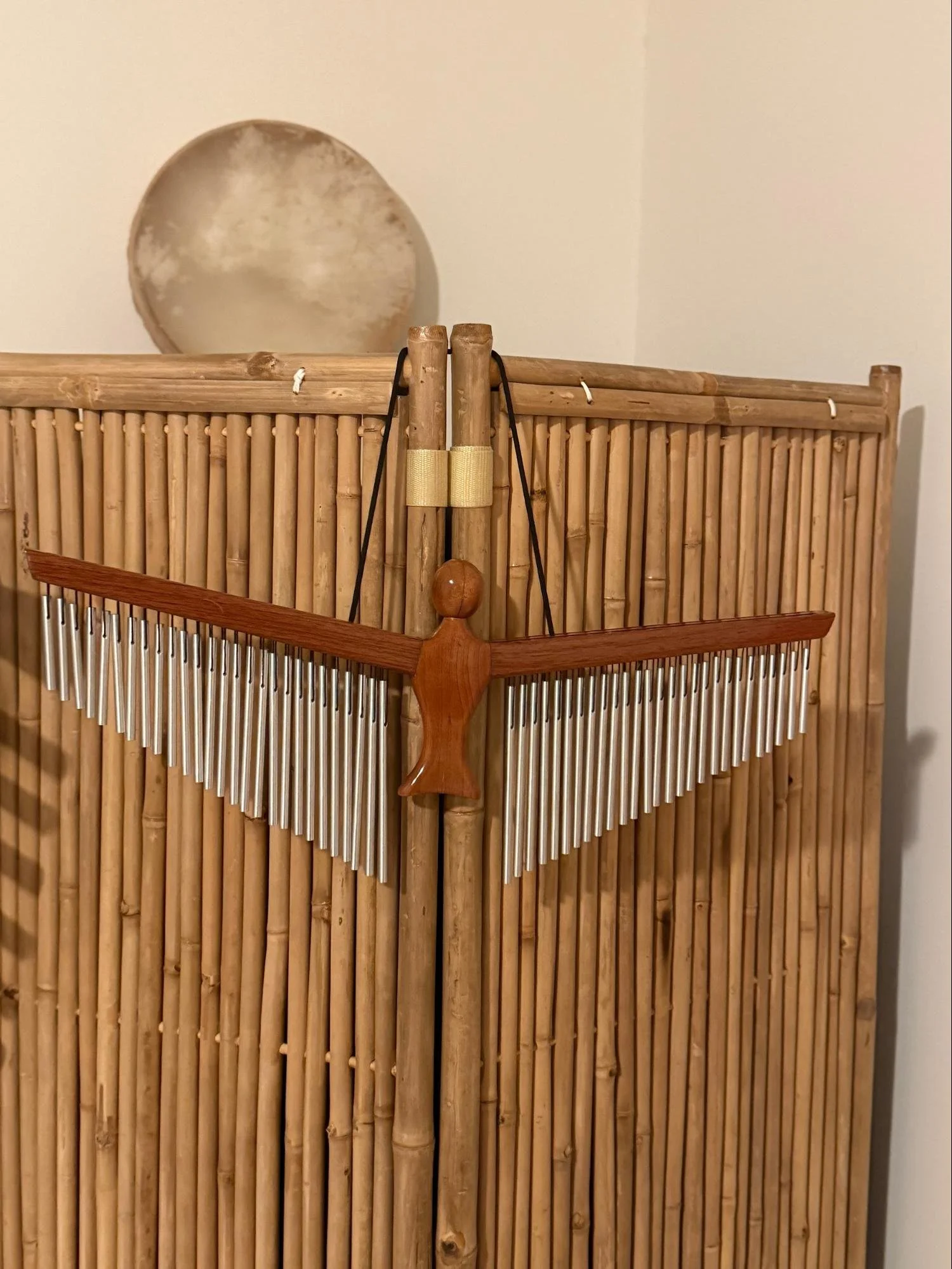 A wooden wind chime hanging on a bamboo panel with a handheld drum in the background.