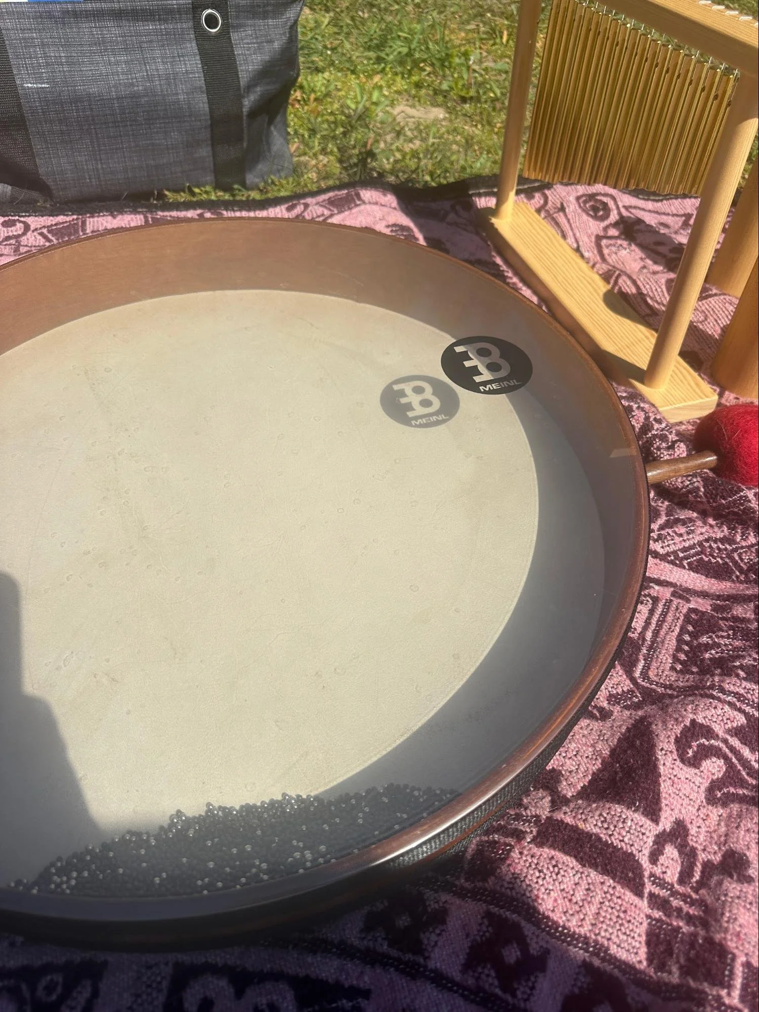 Close-up of a round tambourine with black beads at the bottom, placed on a purple patterned fabric. In the background, there is a wooden chair and part of a black bag, outside on grass.