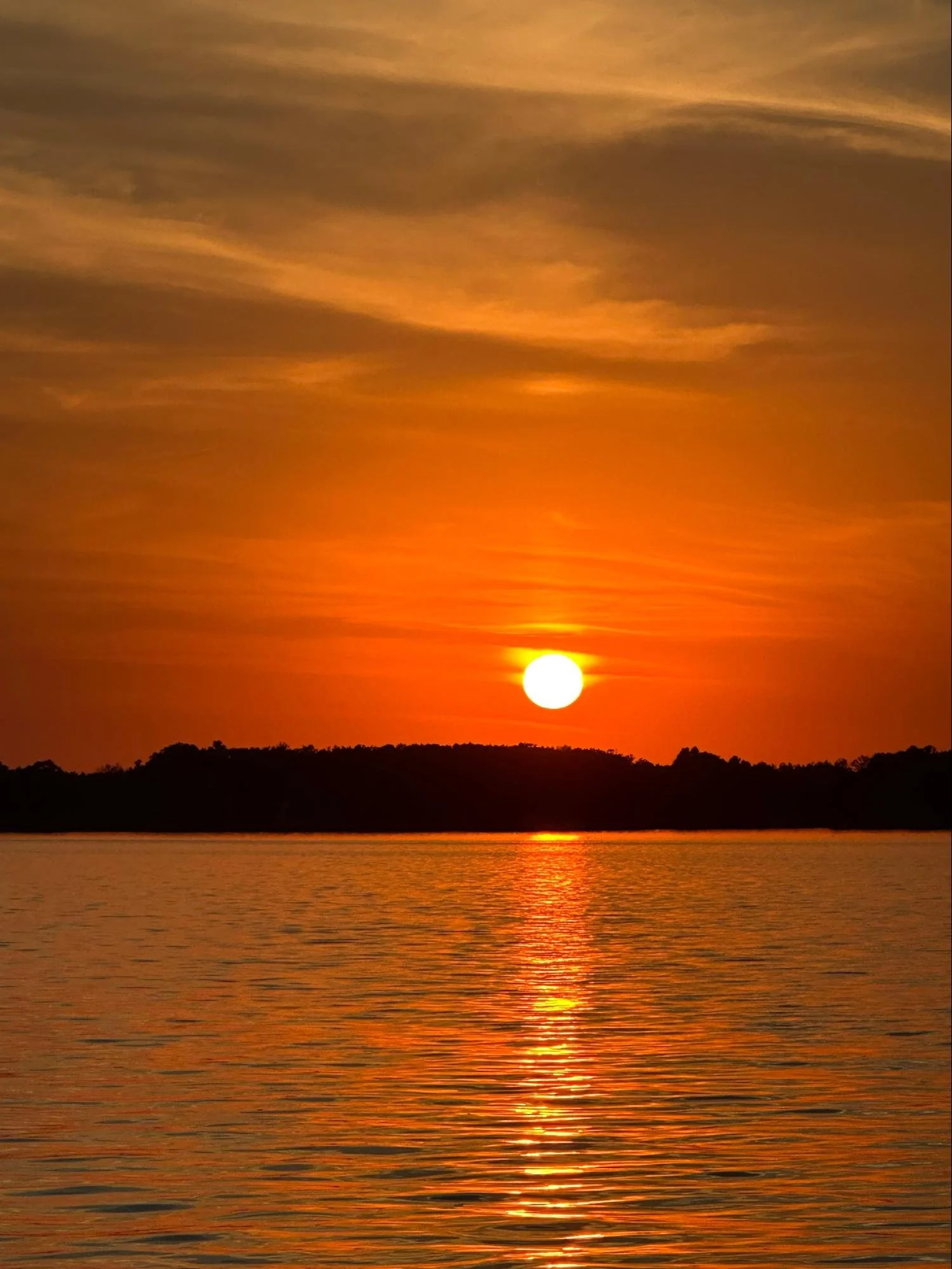 A sunset over a body of water with a silhouette of land on the horizon. The sky is filled with orange, yellow, and pink hues, and the sun is near the horizon reflecting on the water.